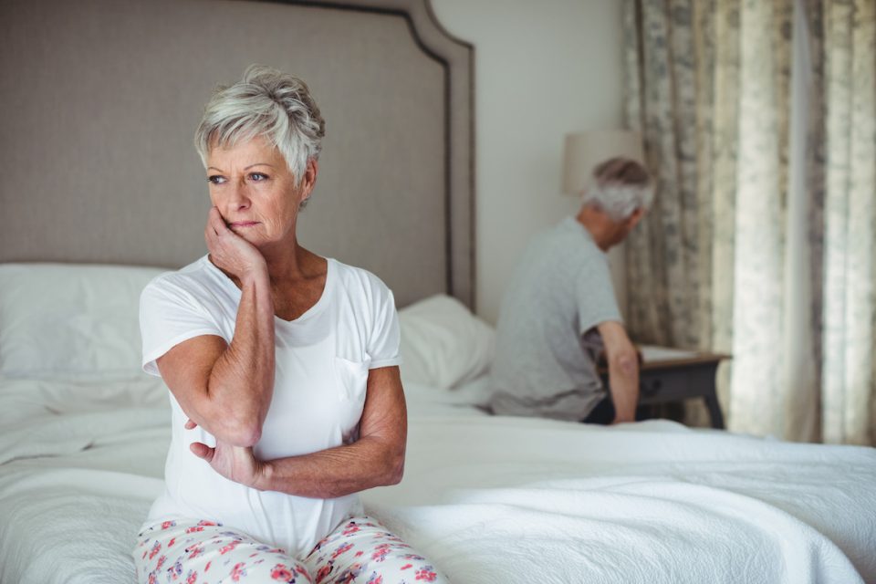 sad senior woman sitting on edge of bend with face in hand. senior man in background sitting on opposite edge of bed
