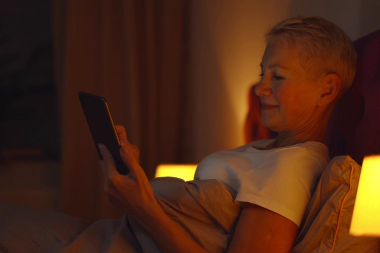 senior woman using smartphone sitting on bed at home in evening