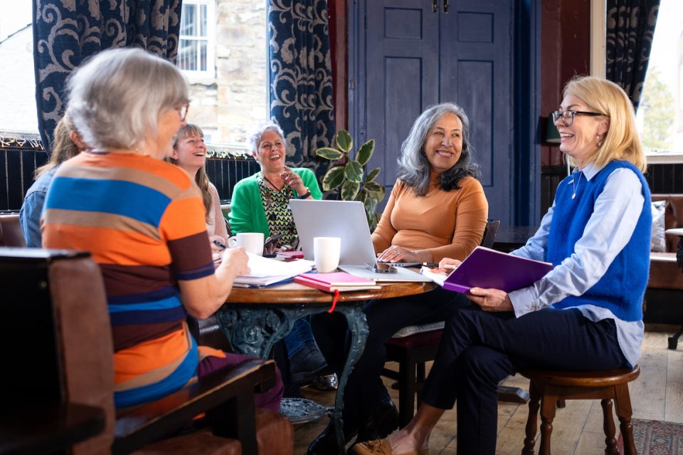 Over-the-shoulder shot of a group of colleagues working from a pub/restaurant. They're having a meeting talking and smiling together to discuss work.