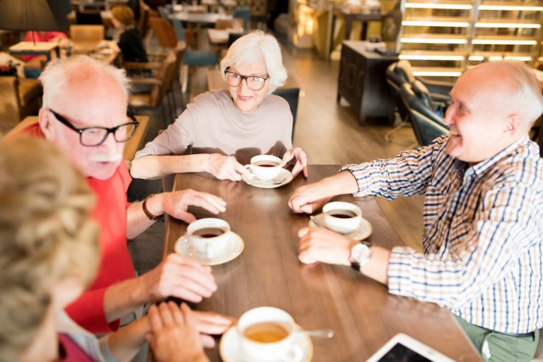 Group of happy senior friends in casual clothing gathering together in cafe to discuss last news and drink tea and coffee