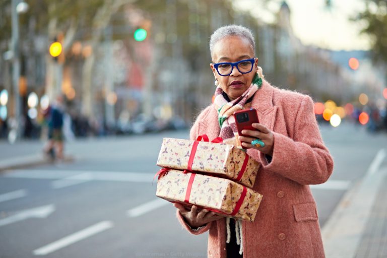 Front view of mature Black woman in winter attire standing curbside with wrapped Christmas gifts and looking at transportation progress report on mobile app.