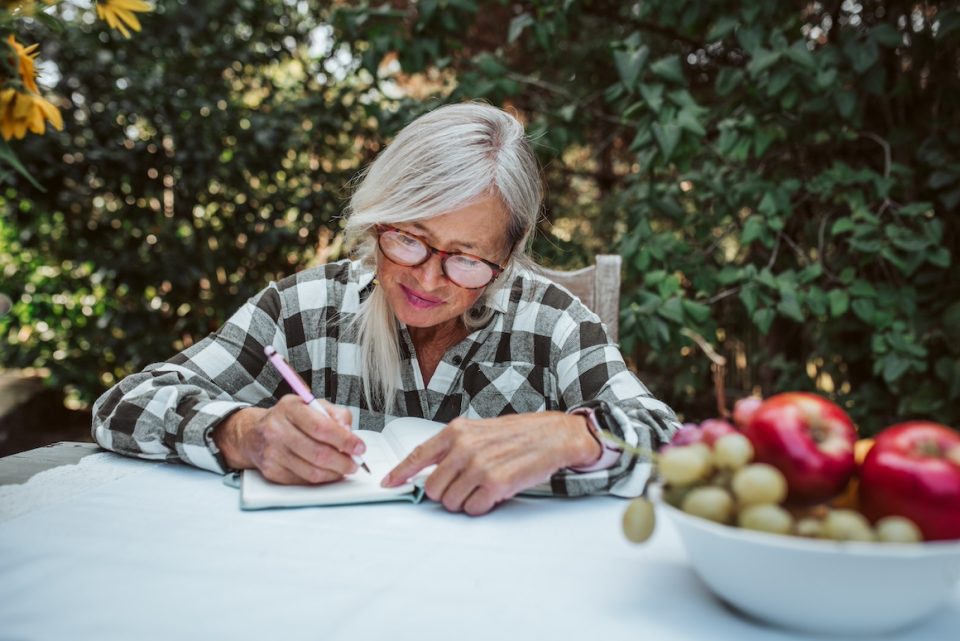 older woman is sitting in the garden and writing in her diary.