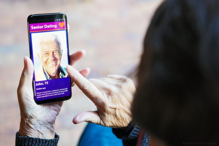 A senior woman's hand nears a mobile phone screen showing a senior man on a seniors' dating app