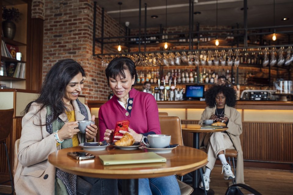 Front view of two mature women sitting side by side in a cafe in Newcastle, England. They are enjoying breakfast and hot drinks in the morning while one woman is using her smartphone to show her friend something. They are on a friends reunion city break for the weekend. One woman is sitting at another table in the background.
