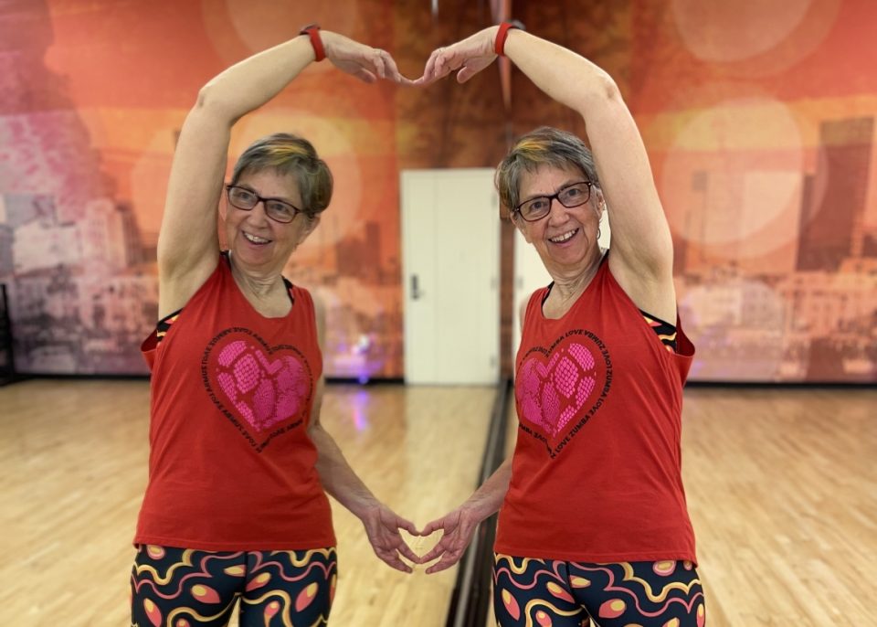 Woman in a exercise studio making a heart symbol with her arms and her reflection in the mirror.