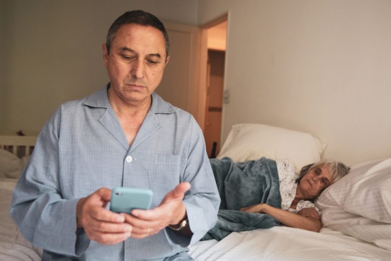Man sitting in bed texting while his wife sleeps.