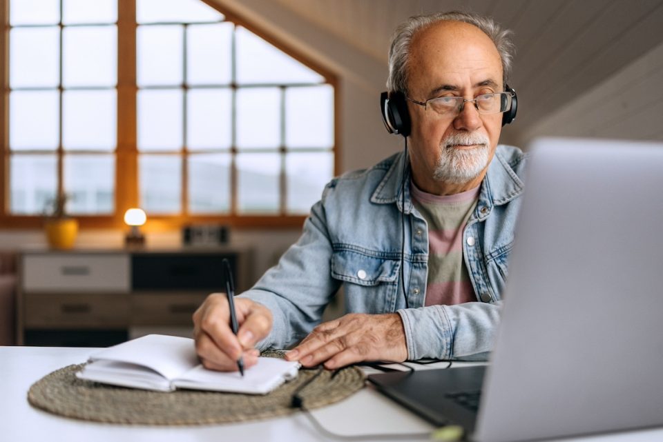 A senior man wearing headphones sits at home, focused on his work while taking notes