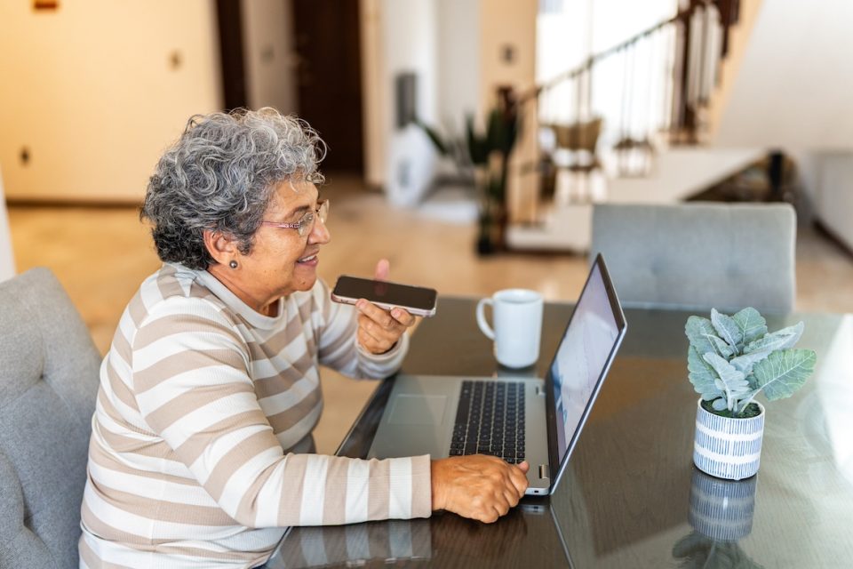 Senior woman using laptop and sending an audio message on mobile phone at home