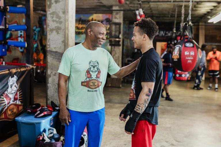 A boxing coach and his athlete share a moment of encouragement and motivation in the gym. The coach places a supportive hand on the boxer's shoulder, reflecting the mentorship and guidance essential in athletic training.