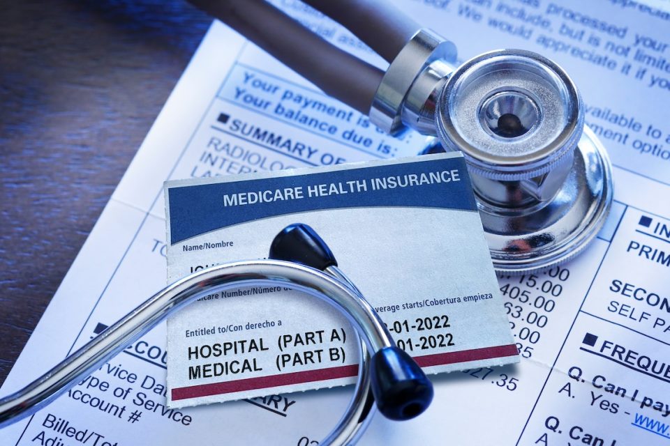 A still life image of a Medicare insurance card resting under a medical stethoscope. The Medicare card sits on top of a medical invoice detailing an array of medical diagnostic test charges.