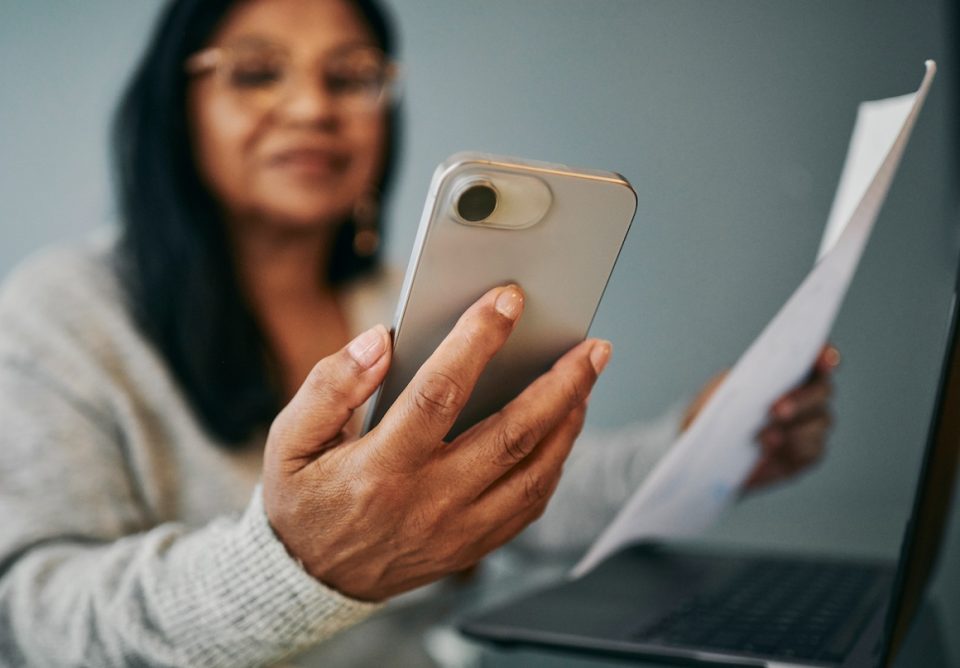 senior woman sits at a desk, reviewing a document and using her mobile phone