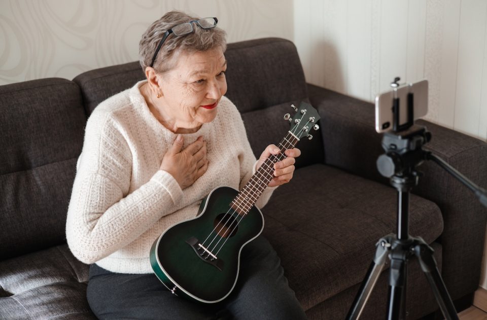 Senior woman with ukulele in her hands, sits on the couch and talks via video chat