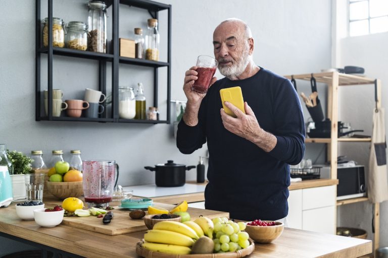 Senior man with smartphone making healthy smoothie at home