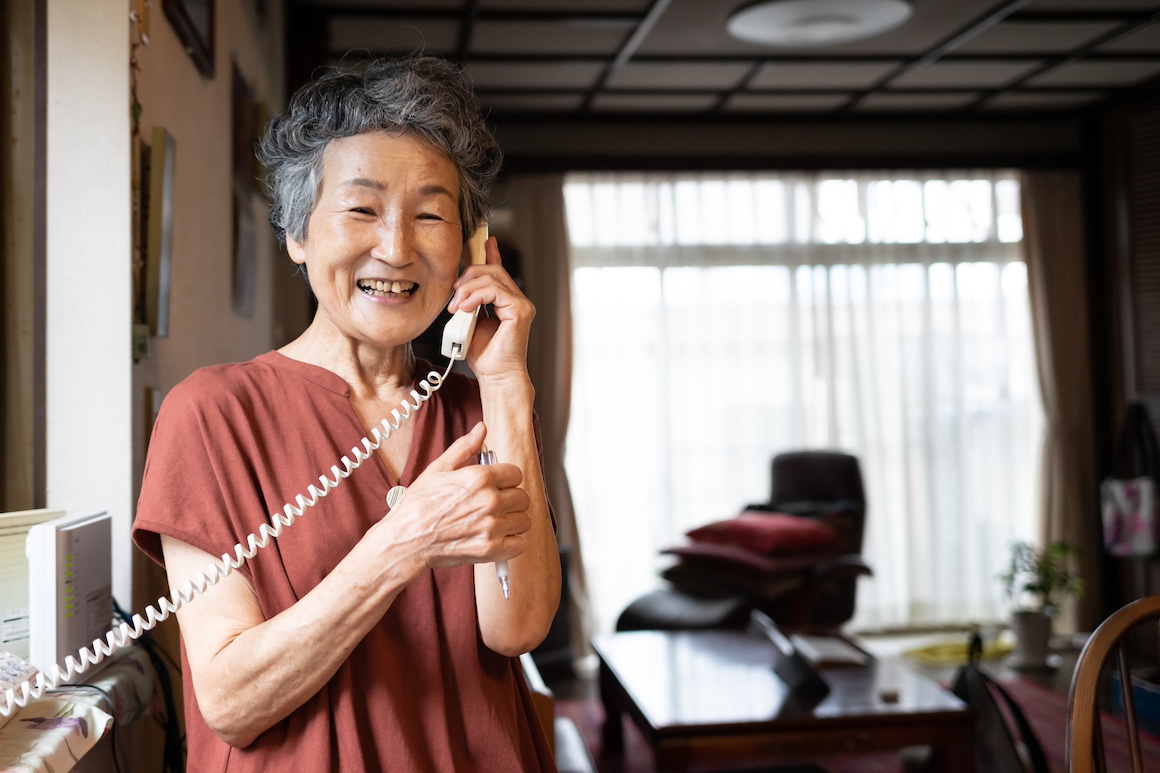 Front view of a senior Japanese woman in her early seventies talking on the landline home phone in the living room.