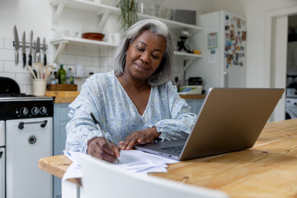 Senior black woman at home writing a letter on paper and using her laptop